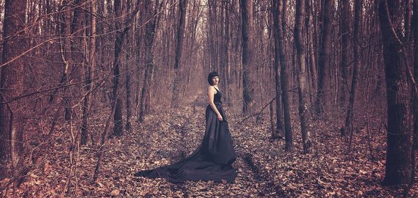 Young woman in black dress standing amidst bare trees in forest