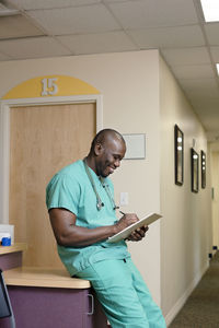 Male doctor writing while leaning on table in corridor