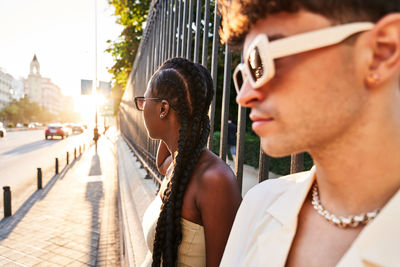 Stylish black woman with braided hair looking at road while leaning on park fence near boyfriend at sundown in city