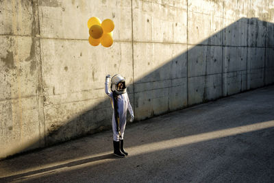 Little girl wearing space helmet holding balloon standing on street against wall