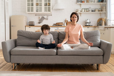 Mom with cute little son doing yoga exercise sit together on sofa at home teaching child to meditate