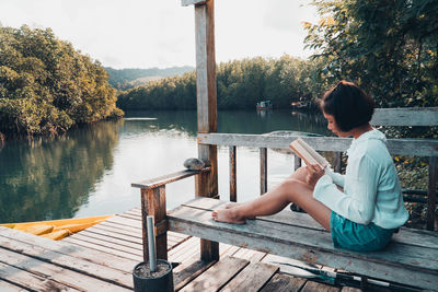 Woman sitting on wood by lake against trees