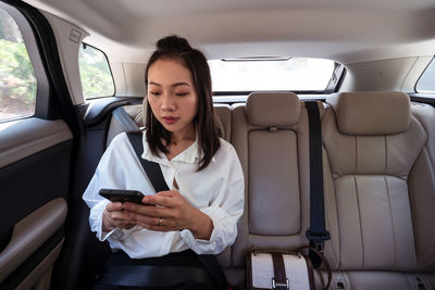 Peaceful ethnic female passenger with fastened seat belt using the cell phone while riding on backseat in taxi