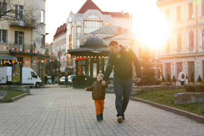 A happy three year old son walks hand in hand with his dad in the city on a sunny spring day. 