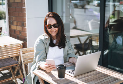 Adult smiling brunette business woman working on laptop using mobile phone in cafe at city 