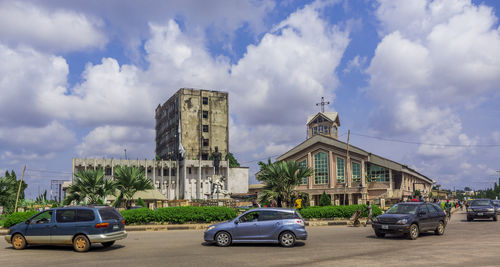 Cars on road by building against cloudy sky