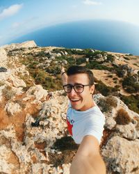 Portrait of happy man taking selfie while standing on cliff against sky