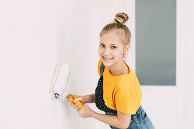 A girl in a denim overalls and a yellow t-shirt helps to paint the walls in an apartment white.