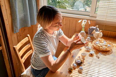Blond woman preparing easter decoration at home, painting colorful easter eggs and coloring egg cups