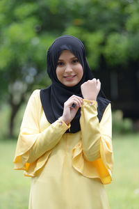 Portrait of smiling young woman standing against trees in park