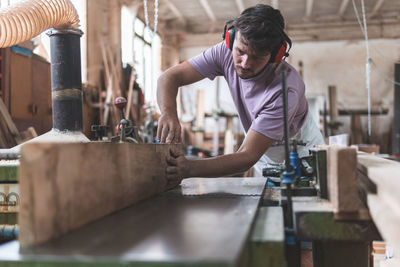 Young male craftsperson holding wooden plank on workbench while working in workshop