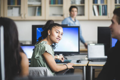 Confident female high school student looking at friend sitting in computer lab