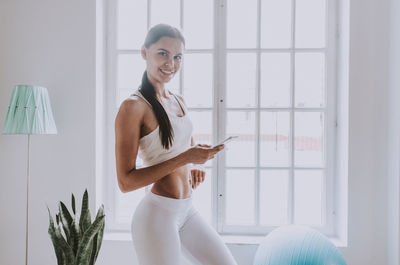 Portrait of smiling woman holding mobile phone standing by window