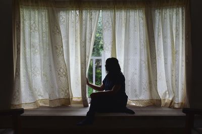 Side view of woman sitting against window at home