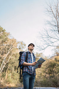 Low angle of male hiker with backpack navigating with paper map while standing in woods and looking away