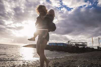 Smiling woman giving piggyback to son at beach against cloudy sky