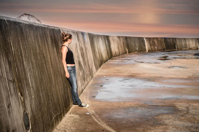 Woman standing by retaining wall against sky during sunset