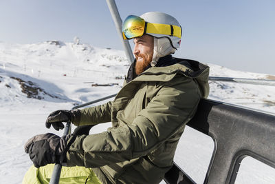 Young man wearing ski helmet and goggles sitting on lift