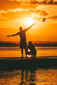 Silhouette of two friends with arms outstretched on beach against sky during sunset