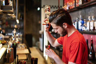 Side view of concentrated bartender pouring alcohol drinking from bottle in metal jigger while preparing cocktail in bar
