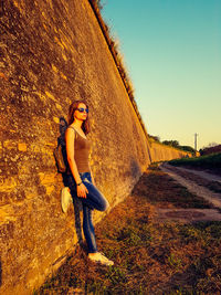 Side view of young woman standing on field by retaining wall against clear sky during sunset