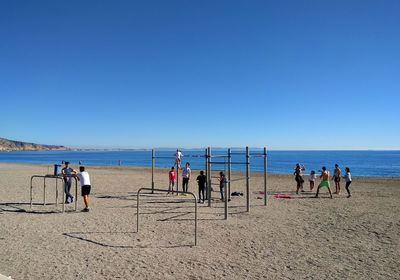 People by play equipment at beach against clear blue sky on sunny day