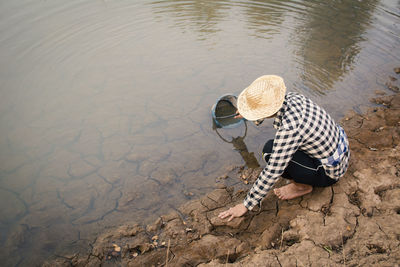 High angle view of person crouching over cracked field while filling water from lake in bucket