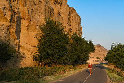 A girl walking on the road below a cliff.