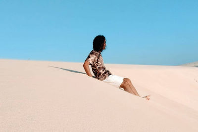 Man sitting on sand at desert against clear sky