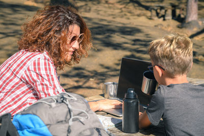 Mother looking at son while using laptop in forest during sunny day