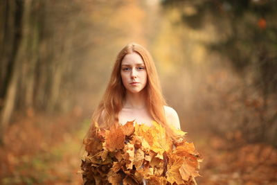 Portrait of young woman covered with leaves standing at forest during autumn