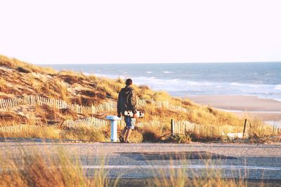 Rear view of man walking with skateboard on street by beach