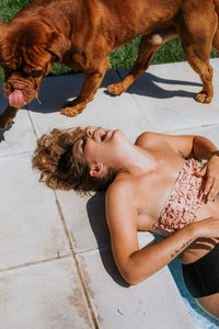 From above of joyful tanned woman in swimsuit laughing while lying on floor and playing with big brown dog on sunny day
