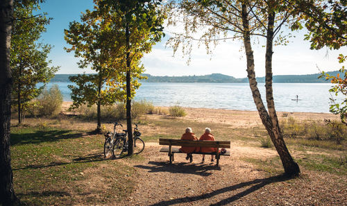 Rear view of people sitting on bench in park by lake