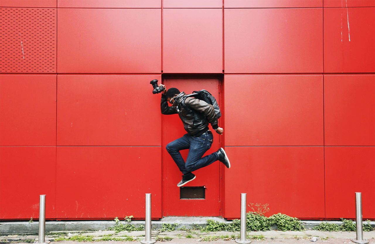 one person, full length, real people, built structure, architecture, wall - building feature, men, casual clothing, lifestyles, building exterior, leisure activity, red, side view, day, jumping, males, outdoors, skill, mid-air