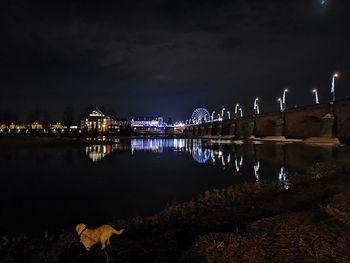 Illuminated buildings by river against sky at night