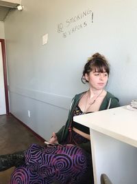 Young woman looking at table while sitting against wall with no smoking sign