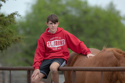 Teenage boy sitting on metallic fence by brown horse at ranch against trees