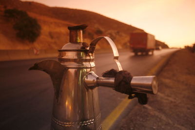 Close-up of metal teapot on road against sky during sunset