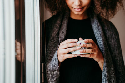 Midsection woman drinking coffee while standing by window at home