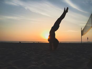 Silhouette woman practicing handstand at beach against sky during sunset