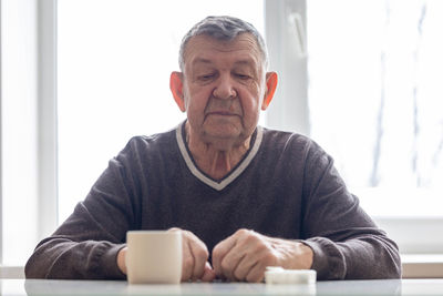 Sad portrait of elderly man. the senior sits at table, thoughtfully looking at mug of water 