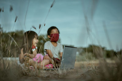 Siblings wearing masks using laptop on land against sky