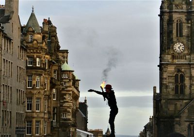 Side view of man with flaming torch gesturing while standing amidst buildings against sky