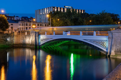 Illuminated bridge over river by buildings against sky at night