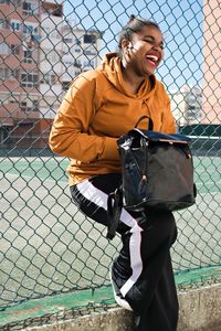 Full length of smiling young woman standing by chainlink fence against sky