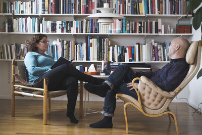 Mature female therapist talking to patient by bookshelf at home office