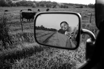 Reflection of boy in side-view mirror leaning out from car window at field