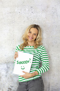 Portrait of smiling beautiful woman holding book with text while standing against wall