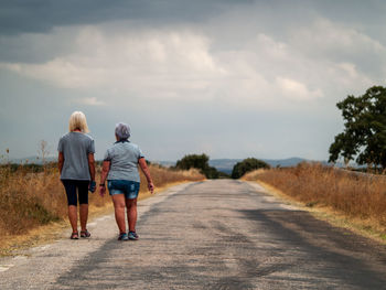 Rear view of female friends walking on road against cloudy sky
